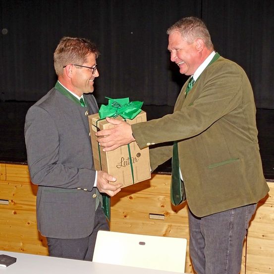 Zwei Männer in Anzügen tauschen eine Geschenkbox aus. Der Mann auf der linken Seite lächelt und hält die Box mit einem grünen Band. Der Mann auf der rechten Seite scheint aufgeregt zu sein, trägt eine grüne Krawatte und einen braunen Anzug. Ein weißer Stuhl steht hinter ihnen.