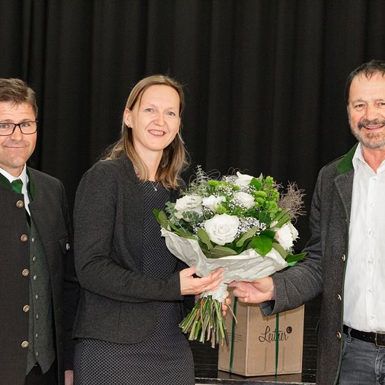 Drei Menschen stehen lächelnd auf einer Bühne. Die Frau hält einen Blumenstrauß. Der Mann rechts trägt einen Gürtel und eine Brille. Hinter ihnen ist ein schwarzer Vorhang und eine Box mit der Aufschrift 'Leuter' zu sehen.