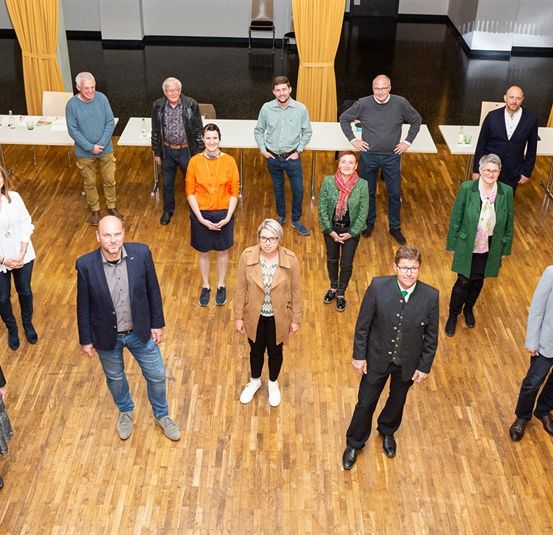 Eine Gruppe von Menschen steht auf einem Holzboden, wahrscheinlich in einem Konferenzsaal. Sie alle schauen zur Kamera. Die Tische und Stühle sind in einer Reihe angeordnet. Der Raum hat einen gelben Vorhang, der an der Wand hängt. Der Boden ist glänzend.