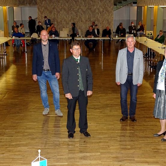 Fünf Personen stehen auf einem Holzboden, möglicherweise bei einer formellen Veranstaltung. Vier Männer und eine Frau sind in formeller Kleidung gekleidet. Sie lächeln und schauen nach vorne. Hinter ihnen stehen lange Tische mit Stühlen und sitzende Personen. Es gibt auch Tische mit Blumen. Der Raum hat Holzwände und einen Holzboden.