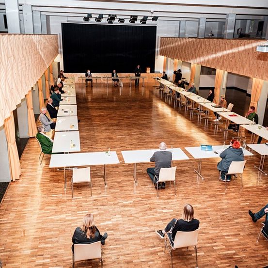 Ein Saal mit Holzböden beherbergt eine Konferenz mit sitzenden Teilnehmern und stehendem Personal im Hintergrund. Tische sind in Reihen angeordnet, und es gibt einen Projektor und Vorhänge an den Seiten.