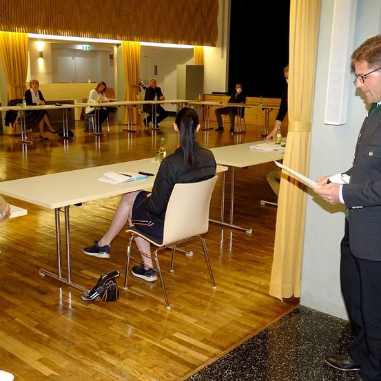 Eine Gruppe von Menschen, die um Tische in einem Konferenzraum herum sitzen, eine Frau mit Rucksack an einem Tisch, ein Mann, der ein Papier liest, und andere im Hintergrund.