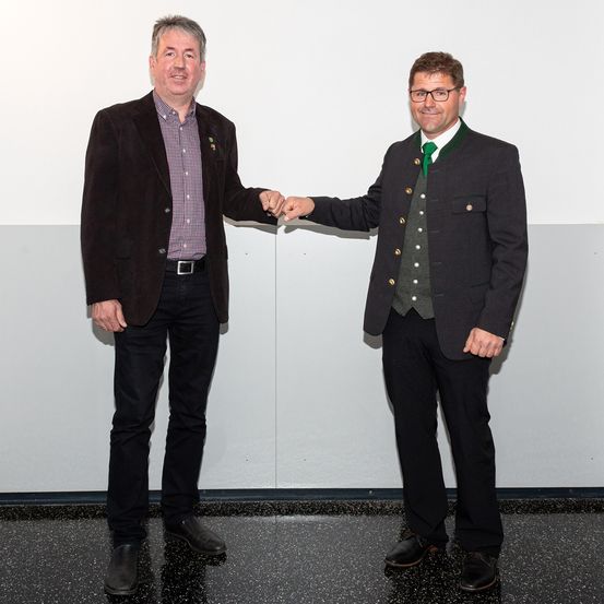 Zwei Männer stehen vor einer weißen Wand und schütteln Hände. Sie sind beide formell gekleidet.