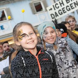 Zwei junge Mädchen mit Gesichtsbemalung stehen in einer Menschenmenge. Eine hat ein Tigergesicht und die andere ein Zebragesicht. Ein Schild im Hintergrund lautet 'Ich bin dagegen'.