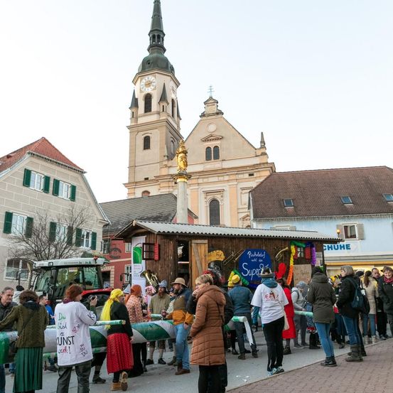 Eine Menschenmenge versammelt sich vor einer Kirche mit Turm und Uhr, in der Nähe eines Traktors. Einige Menschen tragen Kostüme und Hüte, und ein Banner mit einem Musiknoten ist vorhanden.