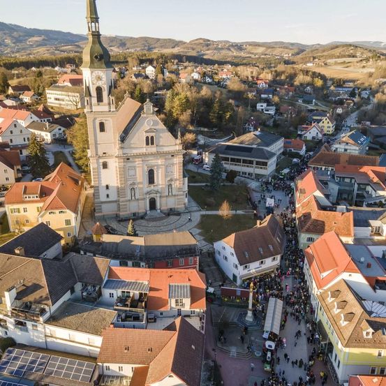 Luftaufnahme einer Stadt mit vielen Gebäuden und einer großen Kirche. Eine Menschenmenge versammelt sich vor der Kirche, während andere durch die Stadt gehen. Die Stadt ist von Hügeln und Bergen umgeben.