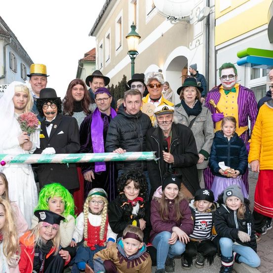Eine Gruppe von Menschen in Kostümen, einschließlich Kinder, posiert für ein Foto vor einem Gebäude mit Straßenlaternen und gelben Wänden.