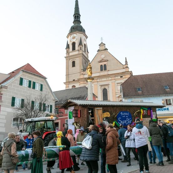 Eine Menschenmenge versammelt sich auf einem Stadtplatz, mit einem Kirchturm und einer Uhr im Hintergrund. Ein Traktor ist in der Nähe geparkt. Einige Leute sind in Kostümen gekleidet und halten Requisiten. Ein kleines Gebäude mit einem blauen Schild steht im Vordergrund.