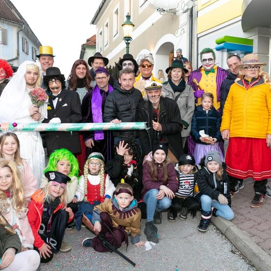 Eine große Gruppe von Menschen in verschiedenen Kostümen posiert für ein Foto im Freien, mit Gebäuden und Straßenlampen im Hintergrund.