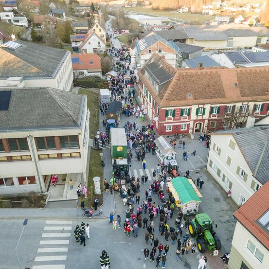 Eine Luftaufnahme einer Stadt zeigt eine Menschenmenge im Zentrum, mit Traktoren und Gebäuden im Blick. Die Stadt hat eine Mischung aus Wohn- und Geschäftsgebäuden, einige mit Solarpanels. Die Menge scheint an einer Parade oder einem Fest teilzunehmen.