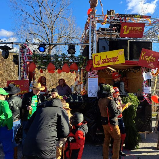Eine lebhafte Veranstaltung im Freien mit Bühne, Dekorationen und versammelten Menschen. Die Bühne hat Lautsprecher, ein gelbes Schild für Gulaschsuppe und rote Schilder für Krapfen. Einige Leute sind in verschiedenen Kostümen gekleidet und halten Luftballons.