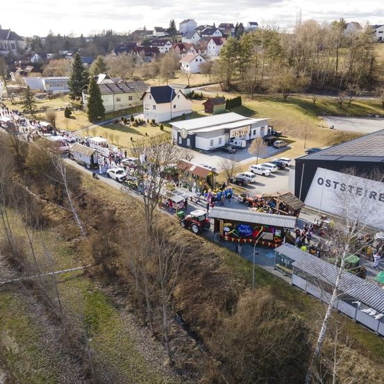 Luftaufnahme einer Parade mit Trucks und Menschen, die auf der Straße gehen, neben Häusern und einem Gebäude mit dem Wort Oststeier darauf.
