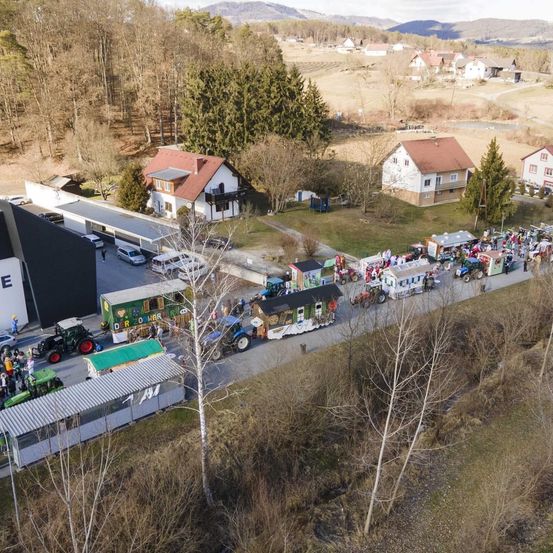 Luftaufnahme einer Parade mit dekorierten Traktoren und Menschen auf einer Straße. In der Nähe sind mehrere Häuser und Autos geparkt, mit Bäumen und Bergen im Hintergrund.