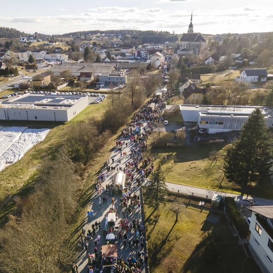Luftaufnahme einer großen Menschenmenge, die sich auf einer Straße in einer Stadt versammelt hat, mit Gebäuden, Bäumen und einer Kirche in der Ferne.
