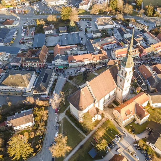 Eine Luftaufnahme einer Stadt mit einer großen Kirche im Zentrum, umgeben von Wohngebäuden und geparkten Autos. Bäume säumen die Straßen und ein Parkplatz ist in der Ferne sichtbar.