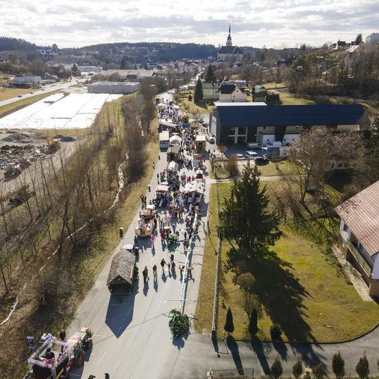 Eine Luftaufnahme einer geschäftigen Straßenmesse mit Menschen, die einen Weg entlanggehen, einem Gebäude mit blauem Dach und mehreren Ständen. In der Ferne befinden sich Bäume, Häuser und ein großes offenes Feld.