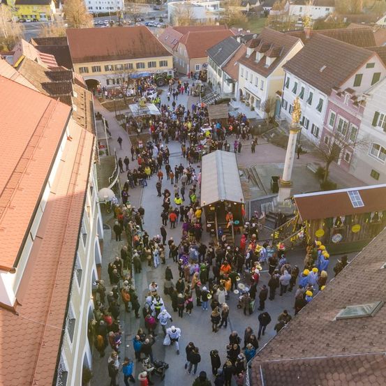Eine Draufsicht auf einen geschäftigen Stadtplatz mit vielen versammelten Menschen, einige in Kostümen, an einem sonnigen Tag. Gebäude mit roten Dächern und vielen Fenstern säumen die Straße.
