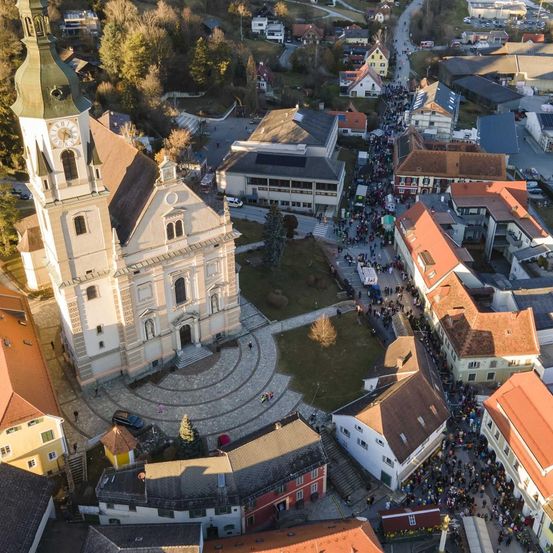 Eine Draufsicht auf eine Kirche mit einem großen Glockenturm und vielen Menschen vor ihr. Die Kirche ist von Wohnhäusern mit verschiedenen Dachfarben umgeben.