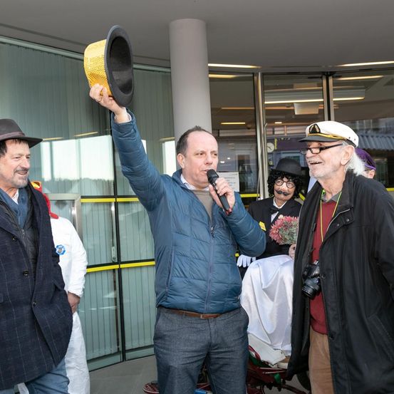 Ein Mann in einer blauen Jacke hält ein Mikrofon und einen gelben Hut und lächelt. Ein anderer Mann mit Hut schaut zu, und eine Frau mit einer Blume steht hinter einem Mann im weißen Anzug.