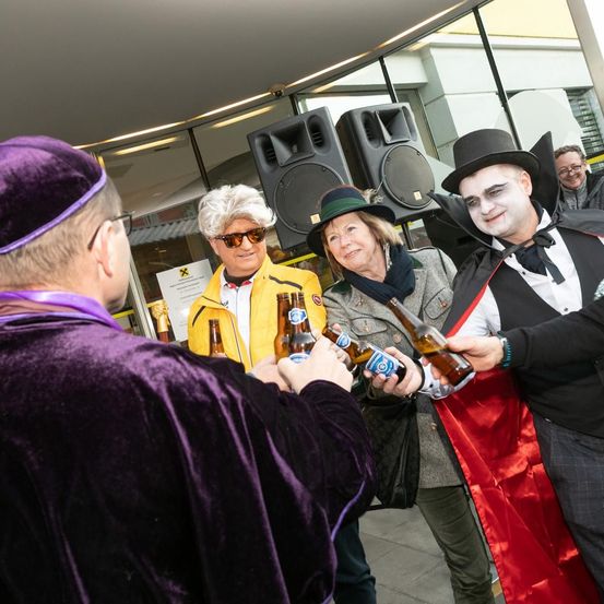 Mehrere Menschen in Kostümen, darunter ein Vampir, eine Hexe und ein Pirat, halten Bierflaschen vor einem Gebäude.