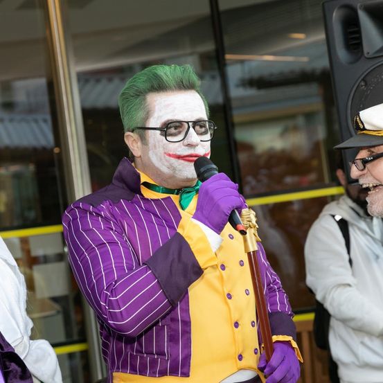 Ein Mann, verkleidet als Joker, spricht in ein Mikrofon und hält einen Stock. Ein anderer Mann in einem Matrosenhut hört zu. Ein Gebäude und ein Lautsprecher sind im Hintergrund.