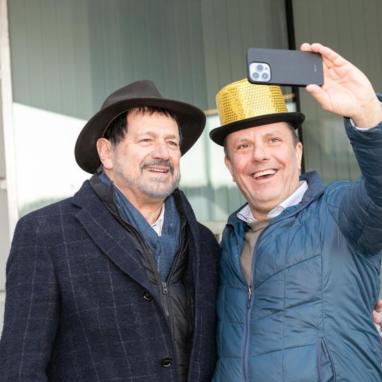 Zwei Männer stehen draußen, einer trägt einen Hut und der andere einen goldglänzenden Paillettenhut, beide lächeln und machen ein Selfie mit einem Handy.