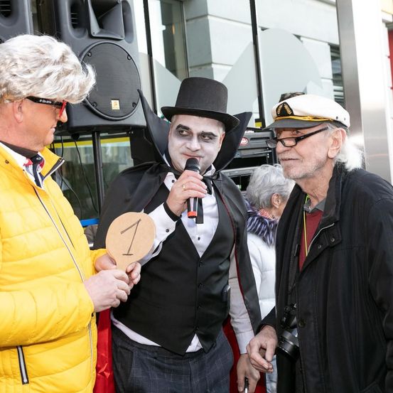 Drei Männer, einer mit einem Mikrofon, stehen vor einem Gebäude. Der Mann in der Mitte trägt einen Dracula-Kostüm, und der Mann auf der linken Seite hält ein Nummer-eins-Schild.