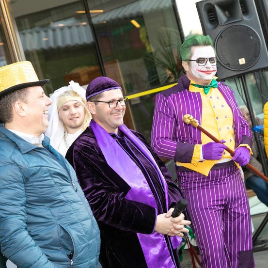 Mehrere Personen sind in Kostümen gekleidet, zwei von ihnen lächeln, einer hält ein Mikrofon. Dahinter steht ein Lautsprecher.