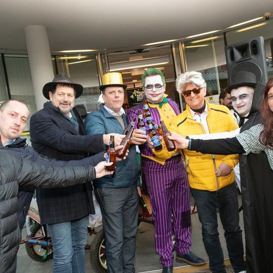 Mehrere Personen stehen zusammen und halten Bierflaschen. Sie sind in verschiedenen Kostümen gekleidet, darunter ein Joker und ein Harlekin. Sie lächeln und posieren für ein Foto.