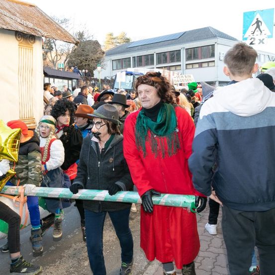 Eine Gruppe von Menschen in Kostümen geht auf der Straße, einige tragen Luftballons. Ein Mann in einem roten Mantel und Schal steht neben einer Frau in einer grauen Jacke. Ein Fußgängerüberweg ist rechts.