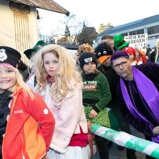 Eine Gruppe von Kindern und Erwachsenen in Kostümen, die draußen stehen. Einige tragen Hüte, einer trägt eine Brille, und ein Schild im Hintergrund lautet 'ICH BIN DAGEEN!'.