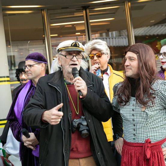 Eine Gruppe von Menschen in Kostümen steht vor einem Gebäude. Ein älterer Mann spricht in ein Mikrofon.