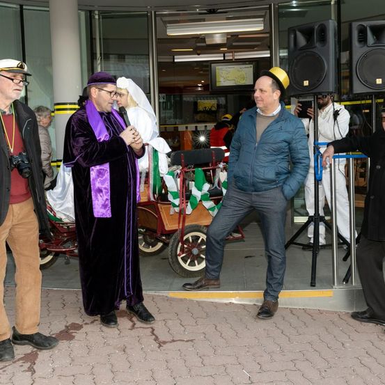 Ein Mann in einer lila Robe spricht in ein Mikrofon, während andere um ihn herum stehen, darunter ein Mann in einer blauen Jacke und jemand, der eine Kamera hält.