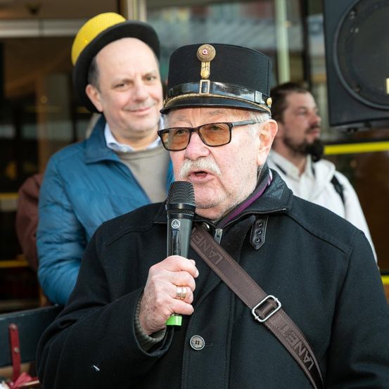 Ein älterer Mann mit Schnurrbart spricht in ein Mikrofon und trägt einen schwarzen Hut mit goldenem Emblem. Hinter ihm schauen ein Mann mit gelbem Hut und ein weiterer Mann in blauer Jacke zu.