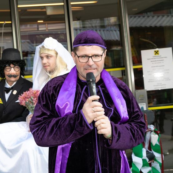 Ein Mann in einem lila Kostüm spricht in ein Mikrofon, während eine Frau in einem weißen Kleid und ein Mann in einem schwarzen Hut hinter ihm stehen.