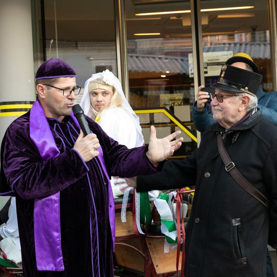 Zwei Männer in Kostümen unterhalten sich vor einem Gebäude. Ein Mann hält ein Mikrofon.