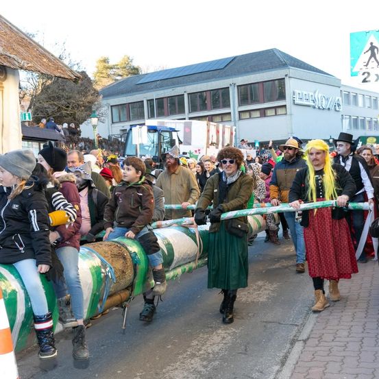 Eine Menschenmenge in Kostümen zieht in einem Umzug und zieht einen langen Baumstamm. Sie sind von Zuschauern und Gebäuden umgeben, eines davon hat ein Schild mit Solarmodulen auf dem Dach.