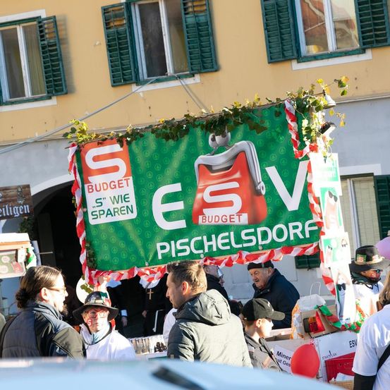 Mehrere Menschen versammeln sich draußen unter einem grünen Banner mit den Texten 'Budget' und 'S' wie sparen'. Sie stehen vor einem Gebäude mit grünen Fensterläden und Pflanzen. Eine Person trägt einen Hut und eine Brille. Es gibt Ballons und Kisten im Bereich.