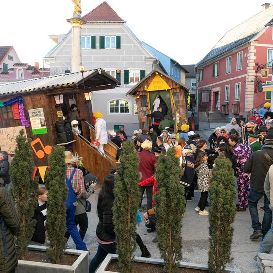 Eine Menschenmenge versammelt sich um eine dekorierte Holzstruktur und eine weitere kleinere Struktur mit einem gelben Schild auf einem Dorfplatz, mit Gebäuden im Hintergrund.