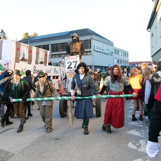 Eine Gruppe von Menschen in Kostümen zieht einen Wagen in einer Parade. Der Wagen hat ein Schild mit der Nummer 27.