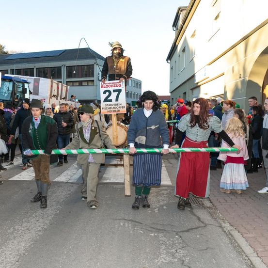 Eine Gruppe von Menschen in Kostümen zieht in einer Parade, wobei sie ein horizontales Banner mit der Nummer 27 tragen. Dahinter steht eine große Figur auf einem Wagen.