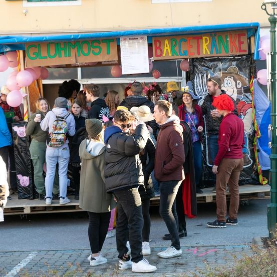 Eine Menschenmenge steht vor einem Stand, der Getränke verkauft. Der Stand hat Luftballons und ein Banner mit der Aufschrift Gluhmost Bargetranke. Einige Leute tragen Kostüme, während andere Getränke in der Hand halten.