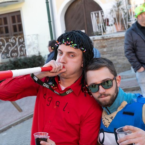 Zwei Männer in Kostümen stehen zusammen und posieren für ein Foto. Einer hält einen Becher und eine Pfeife. Der Hintergrund mit einem Haus und Personen ist unscharf.