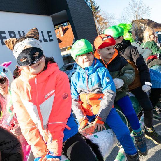 Mehrere Kinder in Kostümen sitzen auf einem großen Baumstamm bei einer Parade. Eines trägt eine Katzenmaske, ein anderes einen grünen Hut und andere rote und blaue Mützen. Ein Gebäude mit dem Wort Frulle ist im Hintergrund zu sehen.