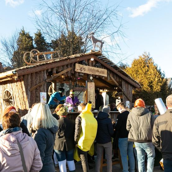 Menschen stehen in einer Schlange vor einem festlichen, hölzernen Stand mit einer Ziegenstatue obenauf. Sie tragen verschiedene Winterkleidung. Eine Person trägt eine Bananen-Kostüm.