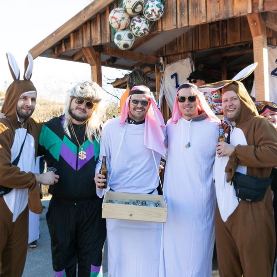 Fünf Männer in Kostümen posieren für ein Foto vor einem hölzernen Gebäude. Jeder hält eine Bierflasche und lächelt in die Kamera. Zwei sind in Hasenkostümen, einer in einem Dollarzeichenhemd und zwei in weißen Roben.