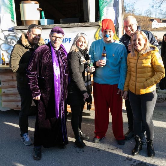 Eine Gruppe von Menschen steht vor einem Imbisswagen, lächelt und posiert für ein Foto. Ein Mann hält eine Bierflasche in der Hand.