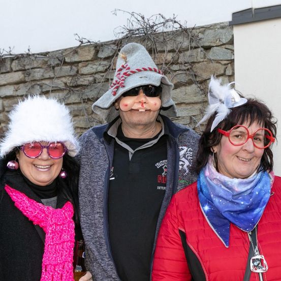 Drei Menschen stehen zusammen, in Winterkleidung und mit Gesichtsbemalung. Die Frau links hält eine Flasche. Hinter ihnen steht eine Steinmauer.