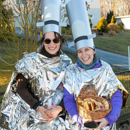 Zwei Frauen in Silberfolie und Kochmützen lächeln, eine hält einen Korb mit Brot. Sie stehen in einem Park mit einem Gebäude und Grünpflanzen im Hintergrund.