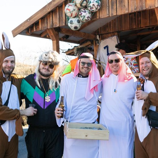 Fünf Männer in Kostümen posieren für ein Foto vor einer Holzstruktur mit hängenden Fußballen. Sie halten Getränke und eine Box.